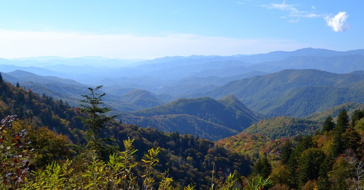 An aerial view of a vast mountain range. The sky is blue and the sun is shining over the trees and foliage-covered mountains.